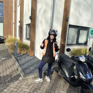 Leerling met helm en oranje veiligheidshesje steekt de duimen op naast een lesscooter met L-plaat